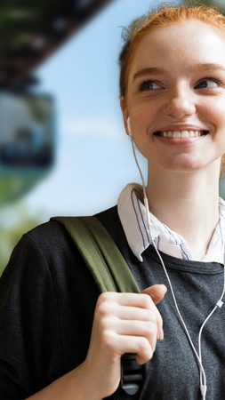 Ein junges Mädchen ist im Fokus des Bildes, sie hat einen Rucksack über eine Schulter gehängt. Im Hintergrund ist verschwommen eine fahrende Schwebebahn zu sehen. 