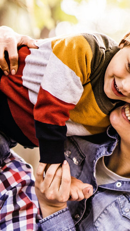 Zu sehen ist eine junge Familie: der Vater hat seinen Sohn auf den Schultern und dieser beugt sich zu seiner Mutter herunter. 